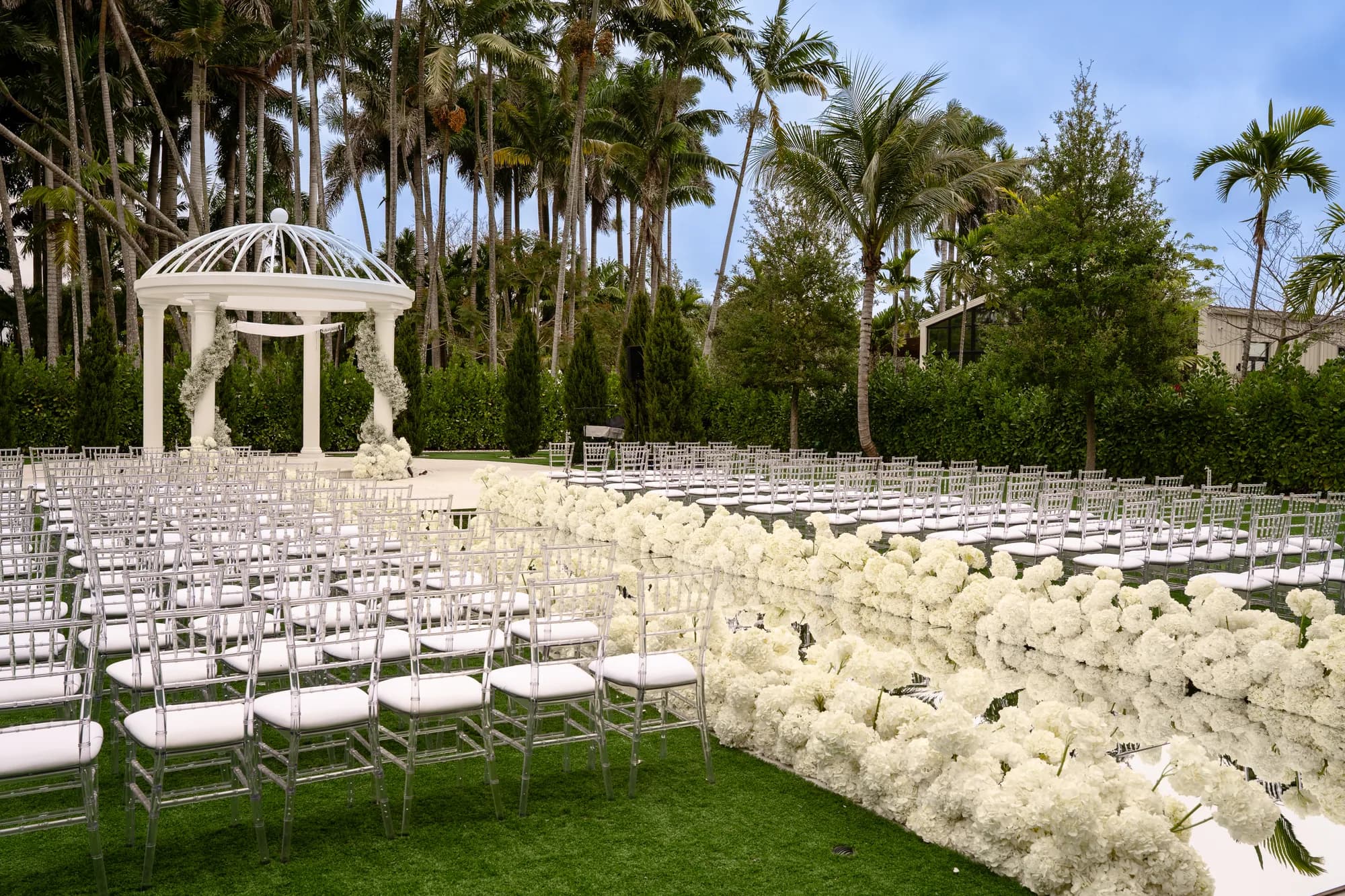 A white wedding pavilion framed by palms, with a white floral aisle running between rows of chairs.