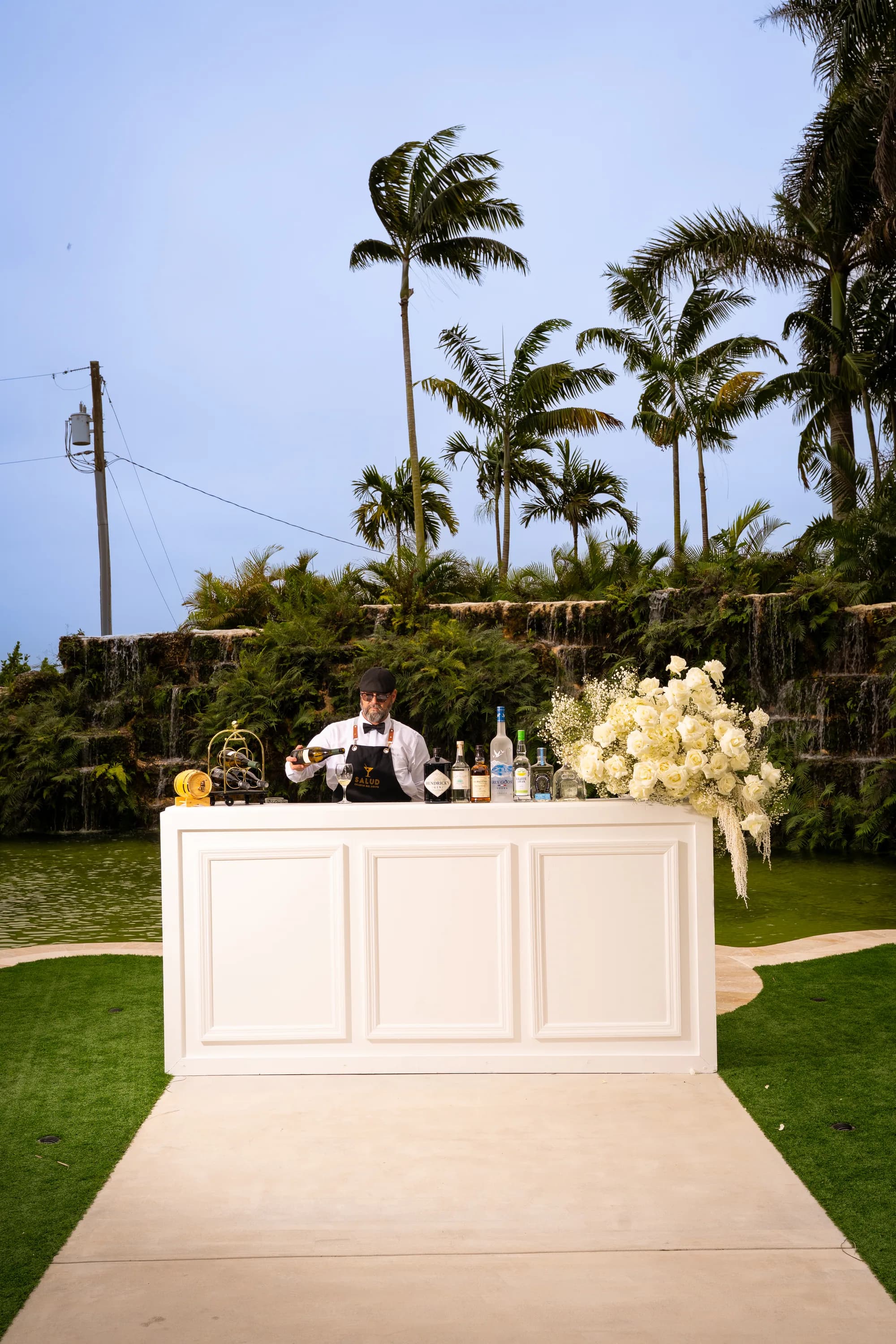 A Salud bartender at the white bar, with spirits and white florals arranged along the top.