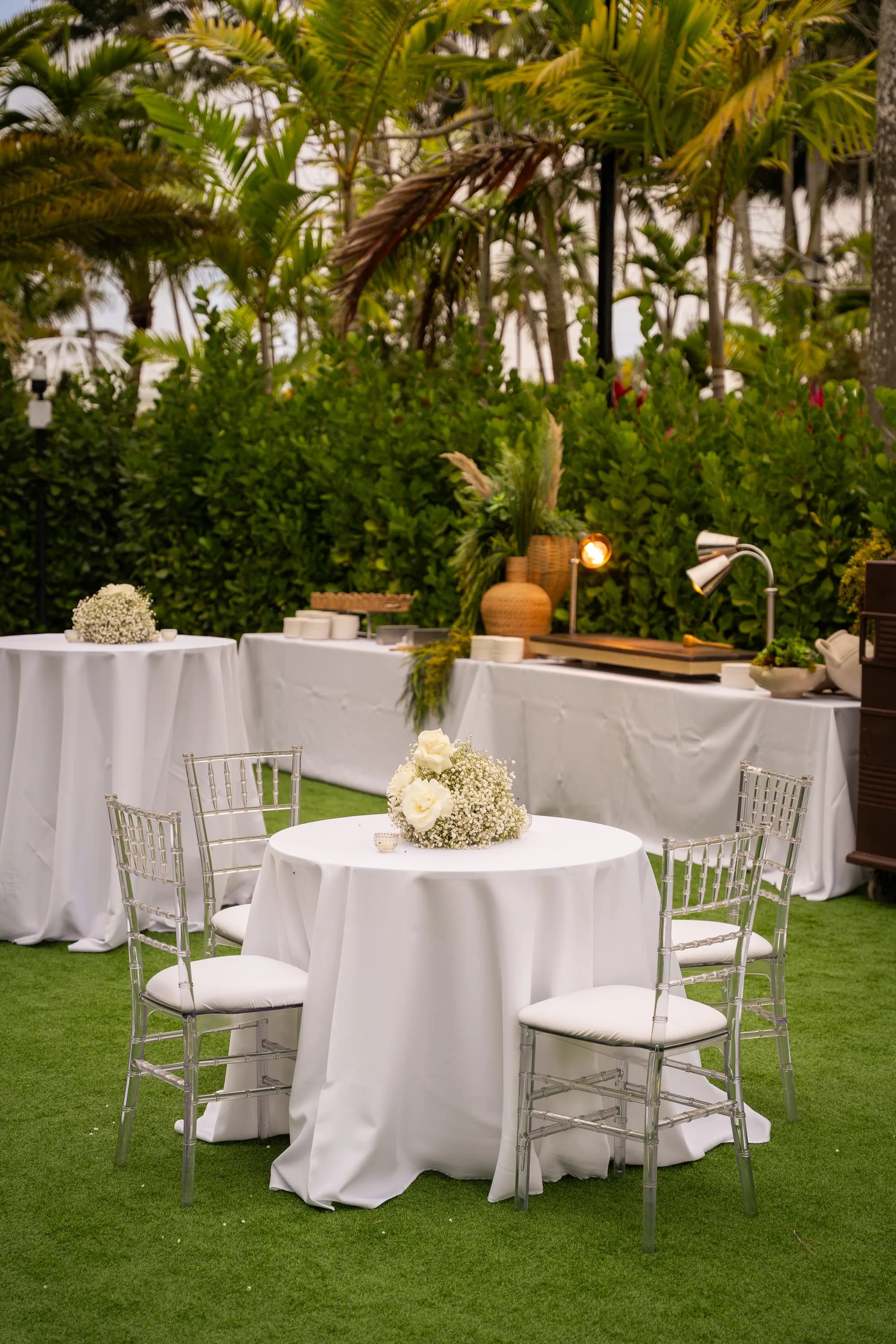 Dressed cocktail tables with white linen and small floral centrepieces on a garden lawn.
